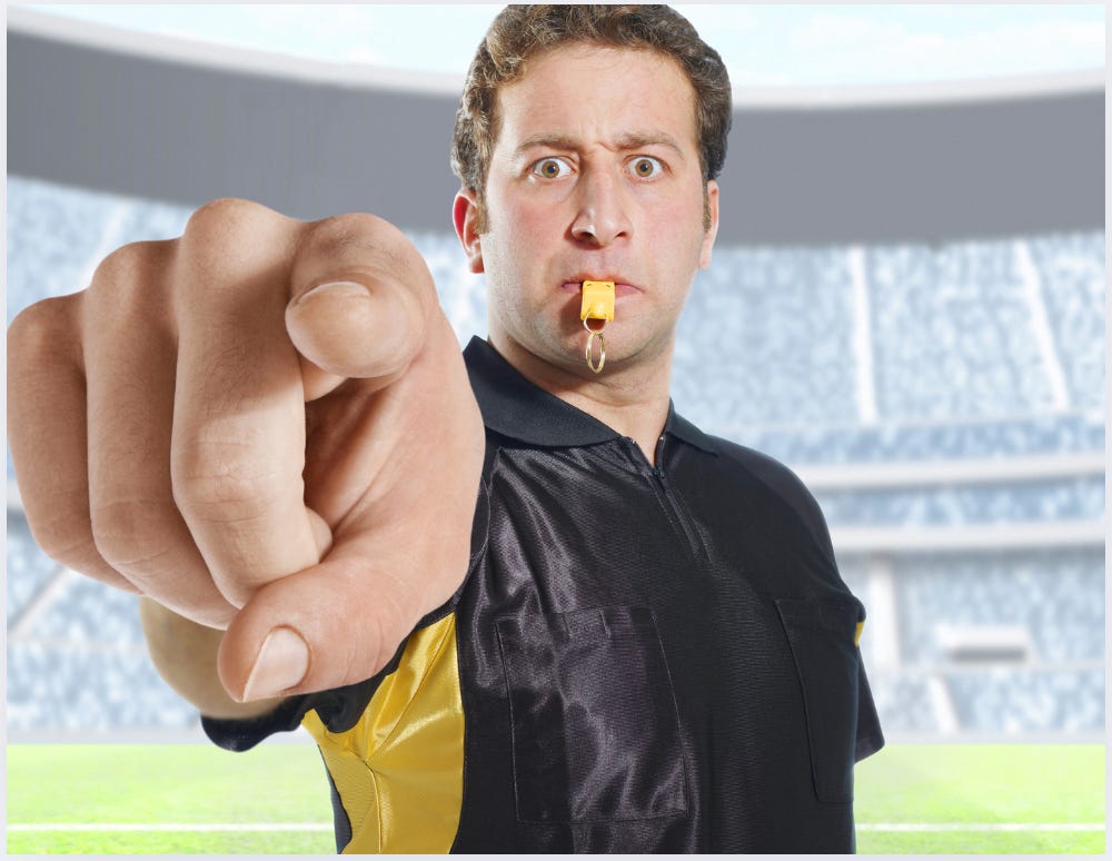Close up image of a referee pointing at the camera with a whistle in his mouth.