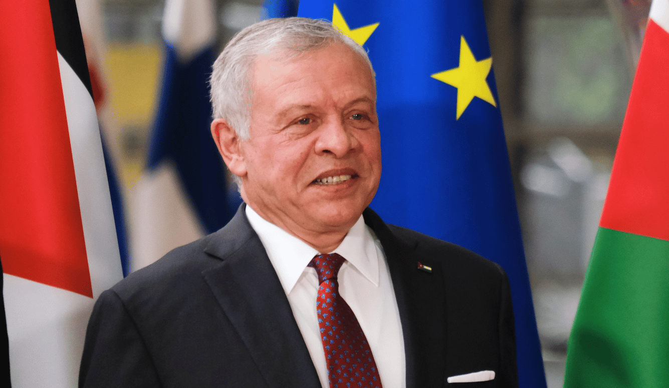 Man in a dark suit and red tie standing before EU and Jordanian flags at a formal diplomatic meeting. Man in a dark suit and red tie standing before EU and Jordanian flags at a formal diplomatic meeting.