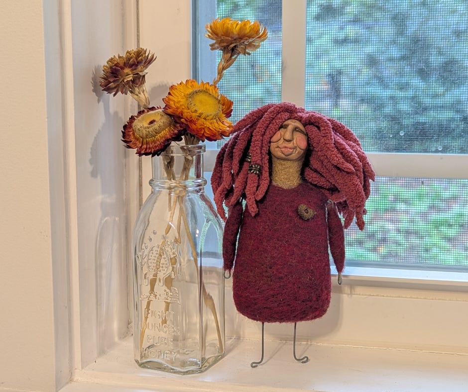 A quirky needle-felted Kitchen Witch doll with red wool hair and a burgundy dress stands on a windowsill beside a glass honey bottle filled with dried orange and yellow flowers.