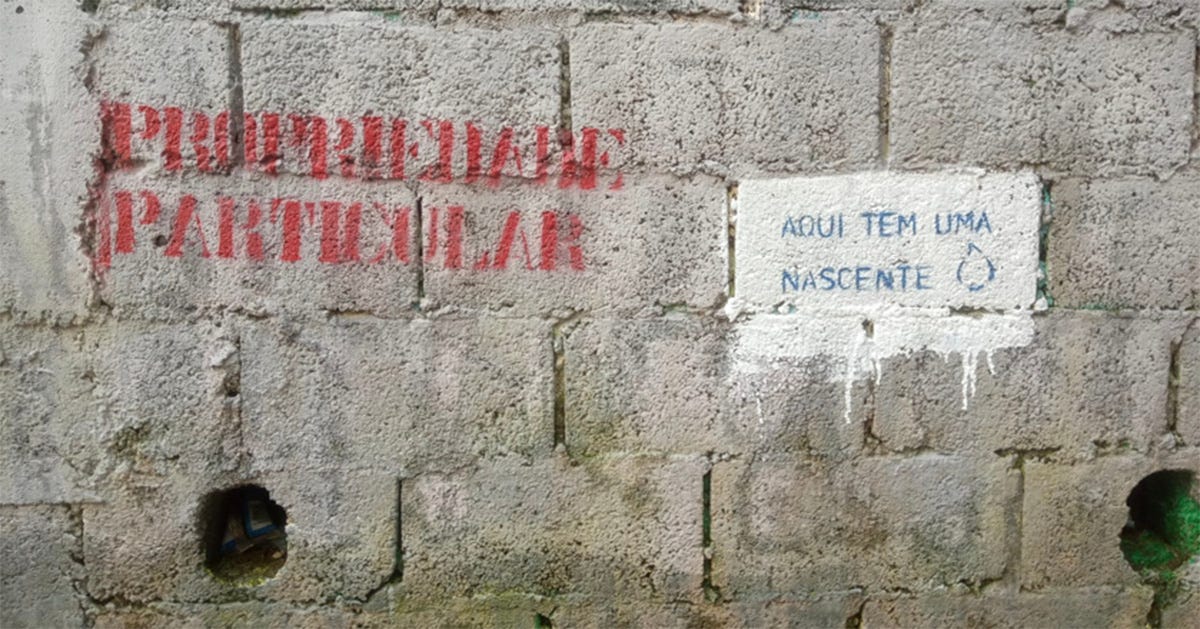 Fotografia de um muro de blocos de concreto com duas intervenções escritas. À esquerda, em letras vermelhas, lê-se “PROPRIEDADE PARTICULAR”; à direita, em uma pequena placa branca, está escrito em azul “AQUI TEM UMA NASCENTE”, acompanhado de um desenho de uma gota. Fotografia de um muro de blocos de concreto com duas intervenções escritas. À esquerda, em letras vermelhas, lê-se “PROPRIEDADE PARTICULAR”; à direita, em uma pequena placa branca, está escrito em azul “AQUI TEM UMA NASCENTE”, acompanhado de um desenho de uma gota.