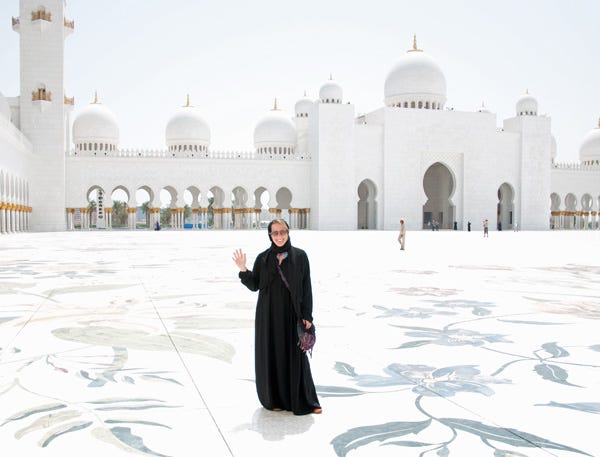 White woman wearing sunglasses and a black abaya robe standing in front of a large white marble mosque.