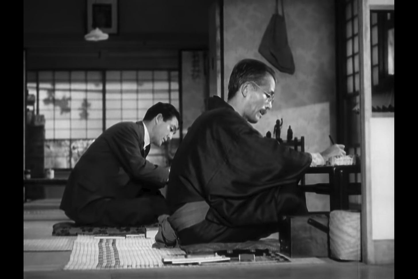 The Weight of Tea: Professor Somiya, wearing a traditional kimono, sits on tatami mats working on Western economics with his assistant Hattori, who wears a Western suit. The Weight of Tea: Professor Somiya, wearing a traditional kimono, sits on tatami mats working on Western economics with his assistant Hattori, who wears a Western suit.