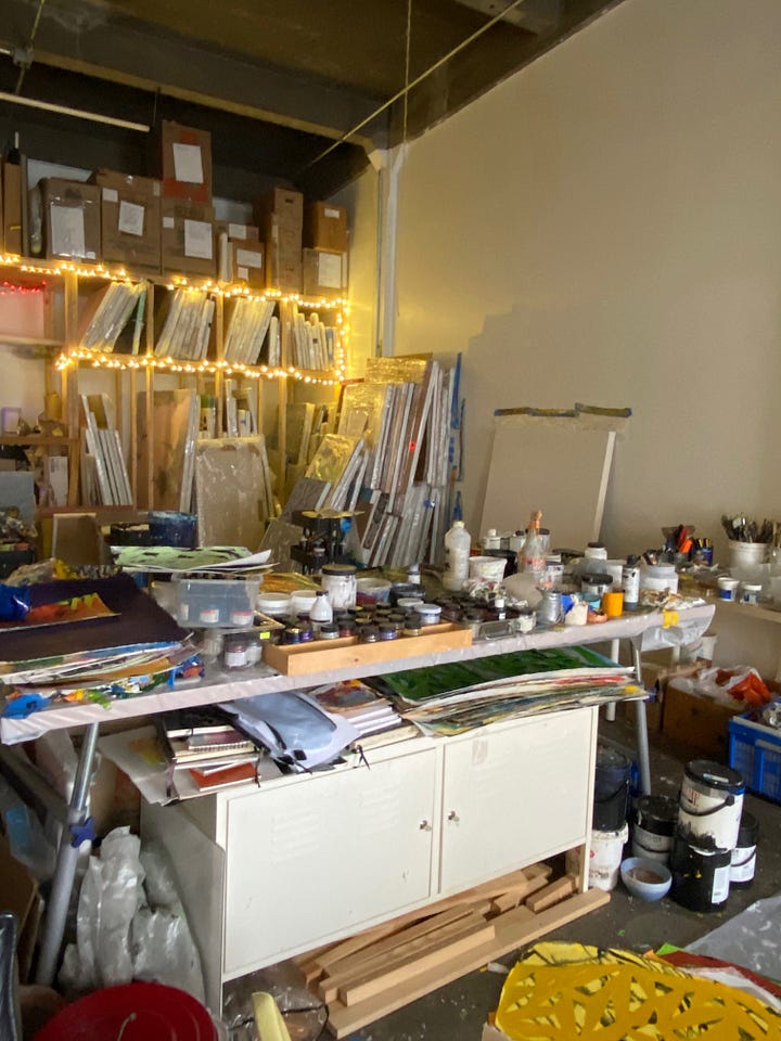 Two photos of my studio space with only natural light coming from a window and from some holiday lights strung across the painting rack.