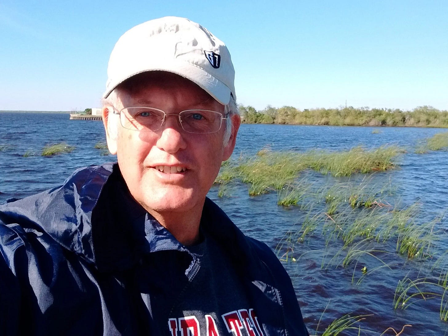 A man wearing a baseball cap and glasses takes a selfie near a windy shoreline with shallow water and tall grasses behind him.