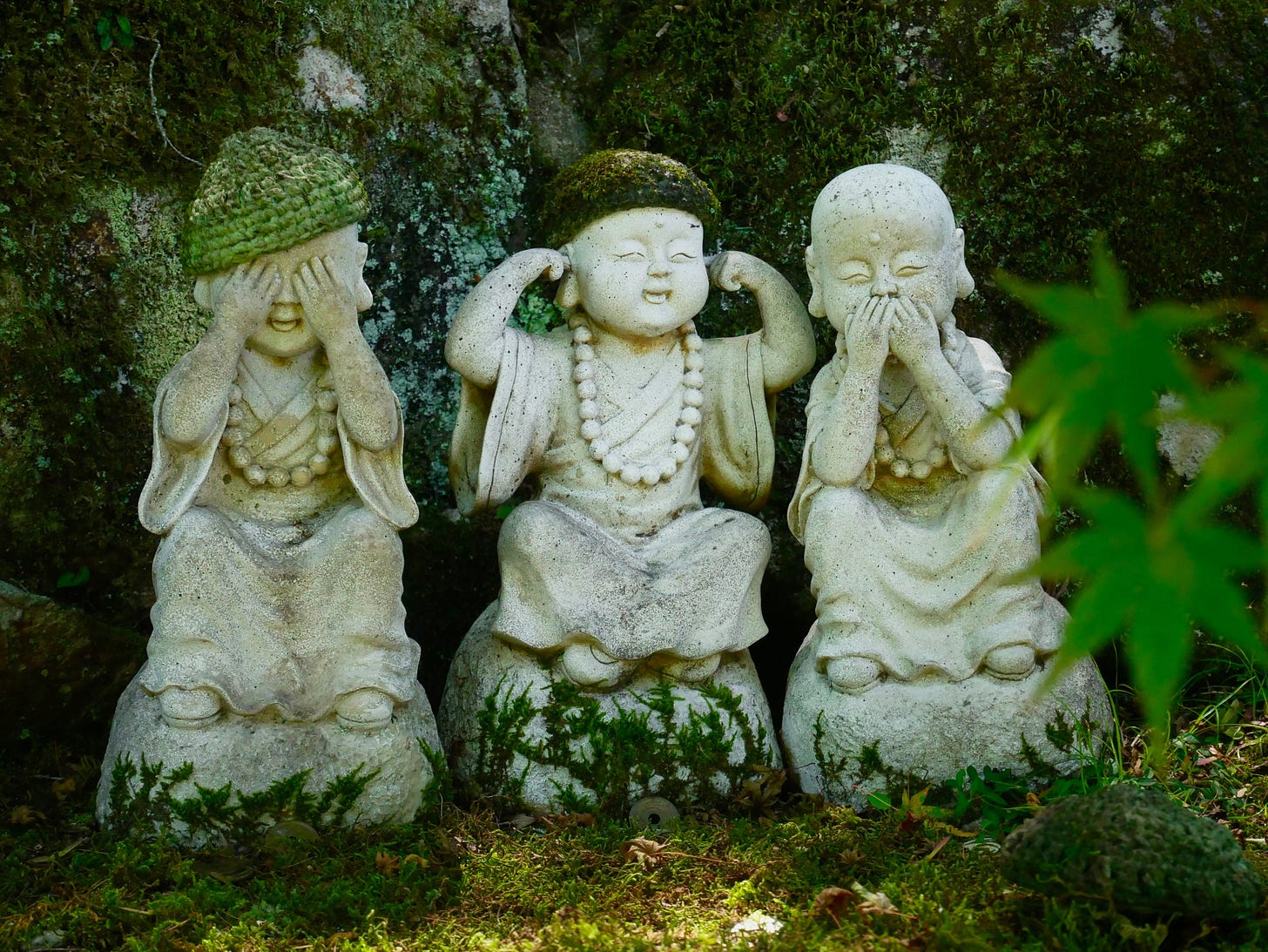 Three weathered stone monk statues sitting side by side against mossy rock, posed as see no evil, hear no evil, speak no evil, with hands over their eyes, ears, and mouth