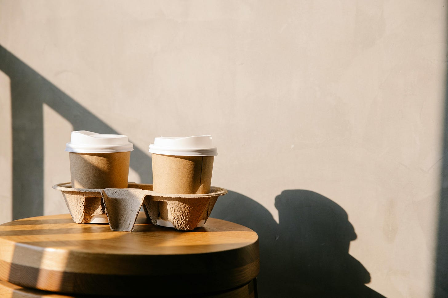 Two brown to go cups of coffee in a to go holder on a small wooden table with shadows, against a tan wall Two brown to go cups of coffee in a to go holder on a small wooden table with shadows, against a tan wall