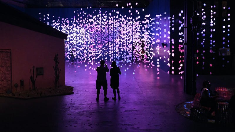Two silhouetted figures stand in the foreground of a dimly lit, cavernous space. With their backs to the camera they are facing a cascade of lights. Strings of colored lights hang down from the high ceiling, creating a magical, suspended “forest” of lights.