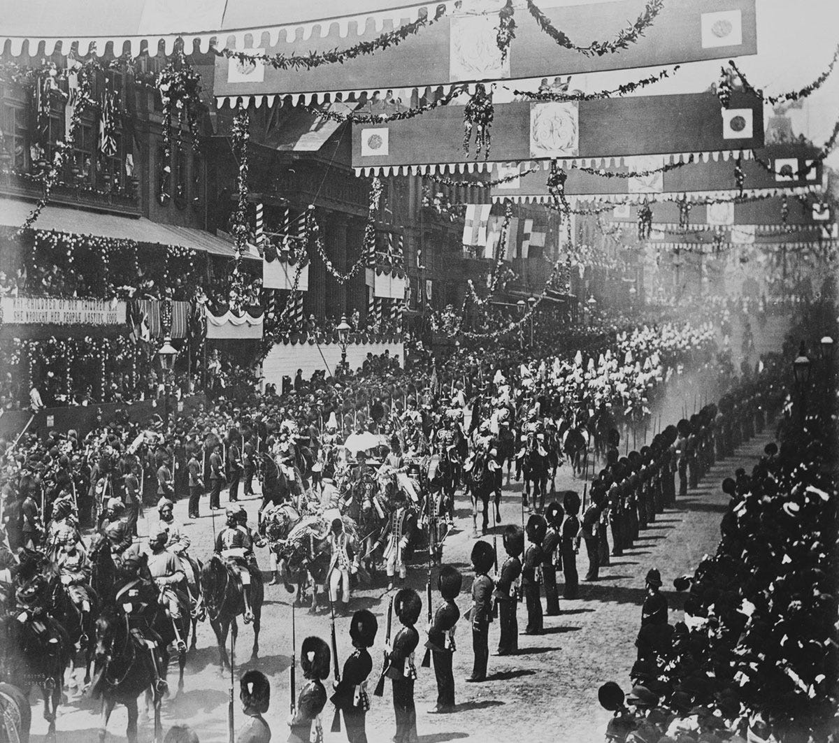 A black and white image of a royal procession. Soldiers and crowds line the street and decorations hang above