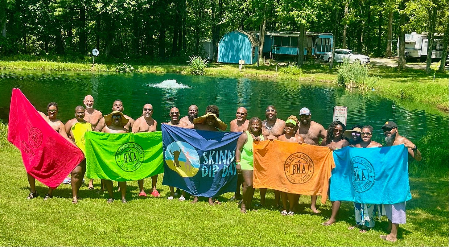 A group of smiling participants with the Black Naturists Association stands on a grassy lawn in front of a pond and fountain, holding colorful BNA and Skinny Dip Day towels. Behind them, trees, cabins, and RVs suggest a naturist campground in Ohio. A group of smiling participants with the Black Naturists Association stands on a grassy lawn in front of a pond and fountain, holding colorful BNA and Skinny Dip Day towels. Behind them, trees, cabins, and RVs suggest a naturist campground in Ohio.