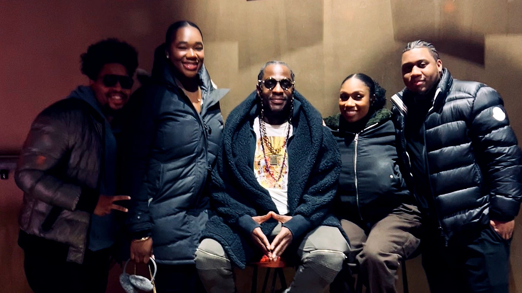 Five of them posed together against a warm-lit wall after the jam: Ray Angry seated at the centre on a stool in a black shearling coat and tinted glasses, layered gold chains over a printed tee, the Juicy sisters and the Council of Goldfinger standing either side in winter coats