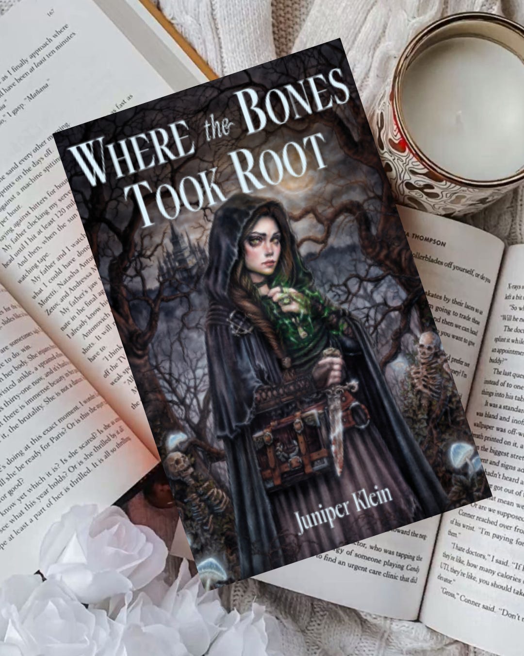 Book "Where the Bones Took Root" on other books next to a candle. Book "Where the Bones Took Root" on other books next to a candle.