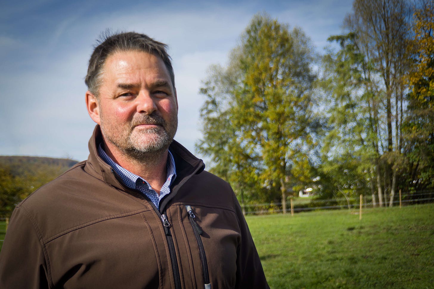 Bio-Landwirt Simon Schönholzer steht auf einer grünen Weide vor herbstlich gefärbten Bäumen. Er trägt eine braune Outdoorjacke über einem karierten Hemd und blickt ruhig in die Kamera. Im Hintergrund ist die hügelige Landschaft seiner Farm zu erkennen. Bio-Landwirt Simon Schönholzer steht auf einer grünen Weide vor herbstlich gefärbten Bäumen. Er trägt eine braune Outdoorjacke über einem karierten Hemd und blickt ruhig in die Kamera. Im Hintergrund ist die hügelige Landschaft seiner Farm zu erkennen.
