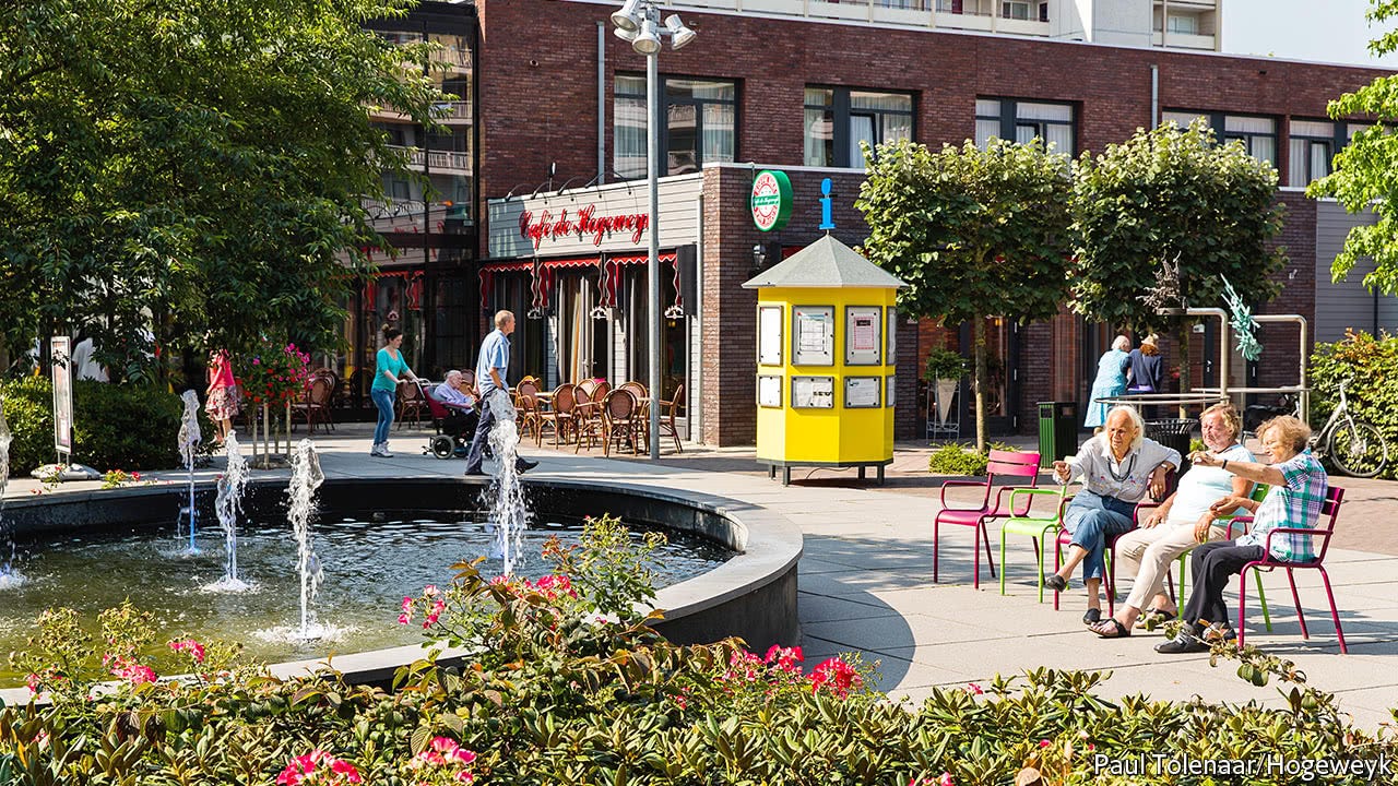 The open-air plaza in the dementia village, with trees and shrubs, large fountain, and tables and chairs for sitting among the cafe, grocery store, and other spaces. The open-air plaza in the dementia village, with trees and shrubs, large fountain, and tables and chairs for sitting among the cafe, grocery store, and other spaces.