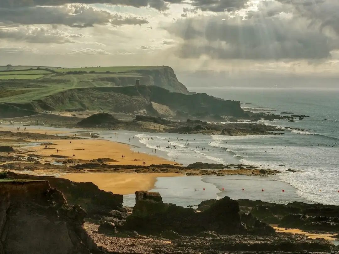A beach sits between a rocky headland in the foreground and a grassy headland behind