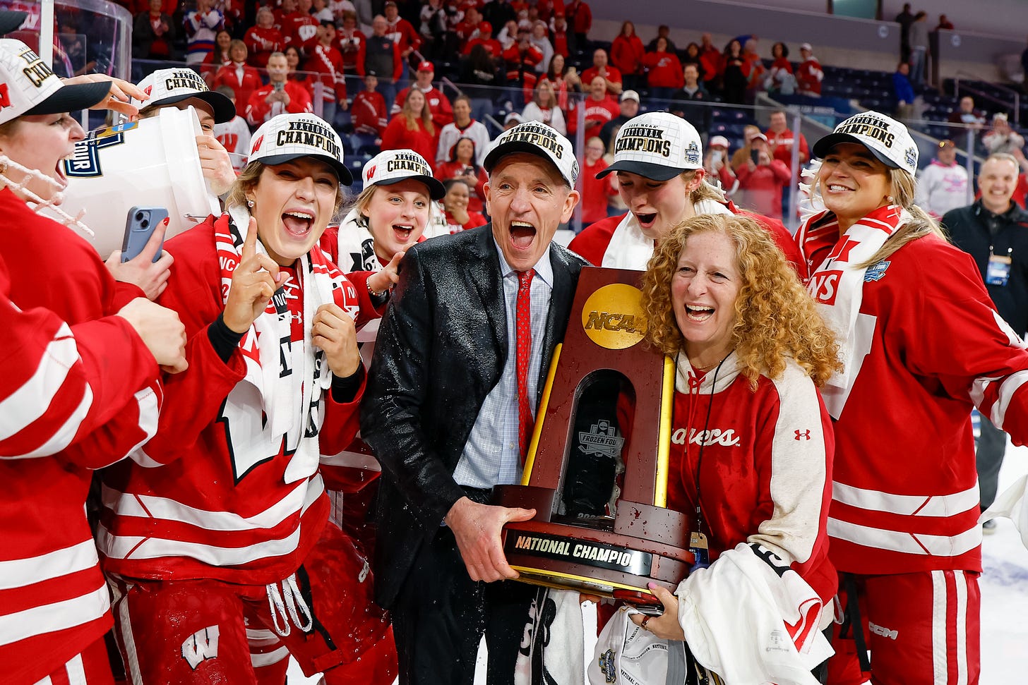 Wisconsin Badgers women's hockey players stand behind Mark Johnson as they pull a bucket out of frame. Johnson and Mnookin hold the national championship trophy together.