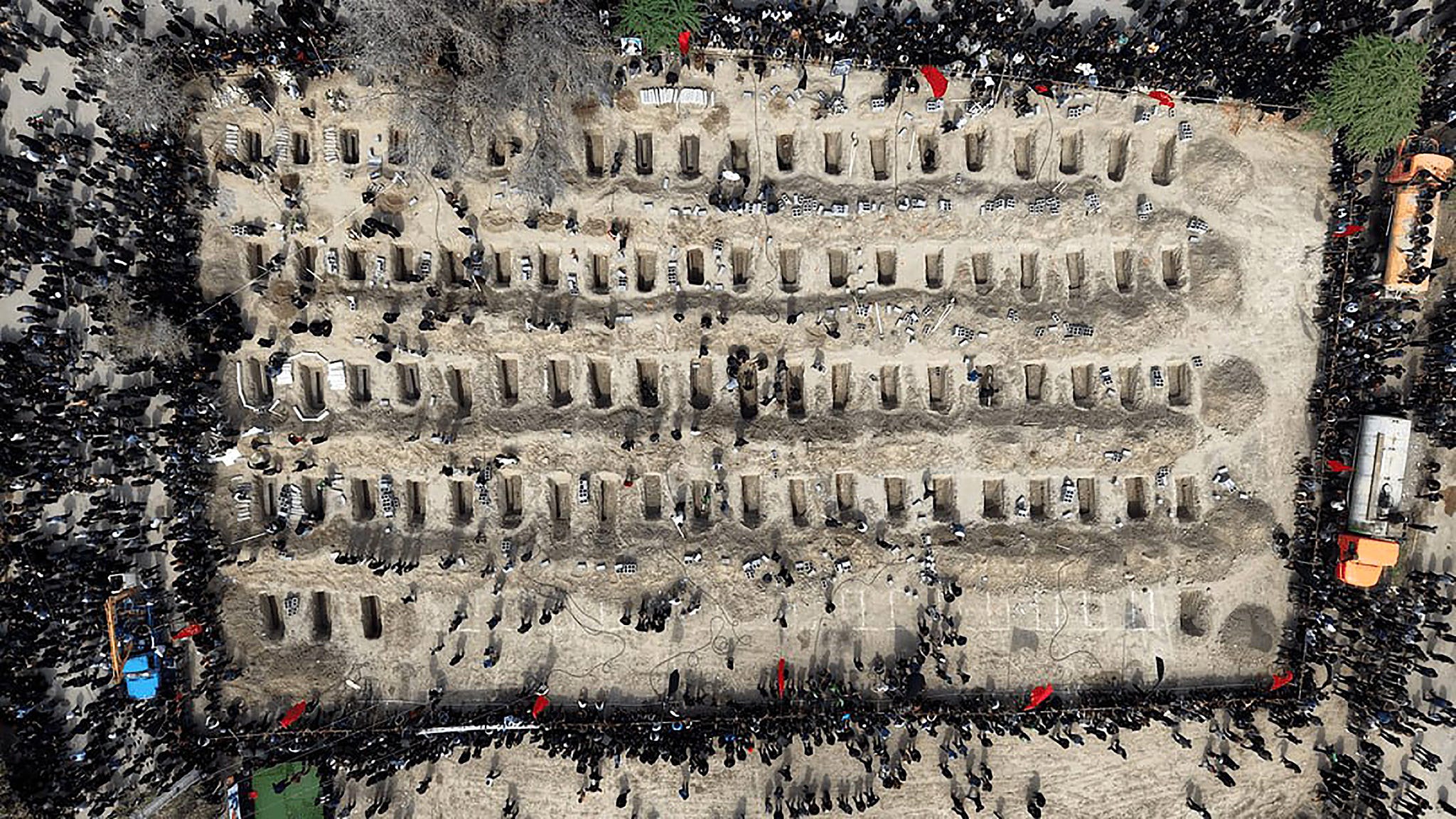 Aerial photograph of a mass burial site in Minab, Iran, showing over a hundred individual graves arranged in long parallel rows across a rectangular excavation. Thousands of mourners dressed in black surround the site on all sides. Several graves are still open, with figures visible inside preparing them for burial. Construction vehicles and red flags are positioned at the edges of the site. The scale of the burial ground, visible only from this overhead angle, conveys the scope of the casualties from the February 28 strike on the Shajareh Tayyebeh girls' elementary school.