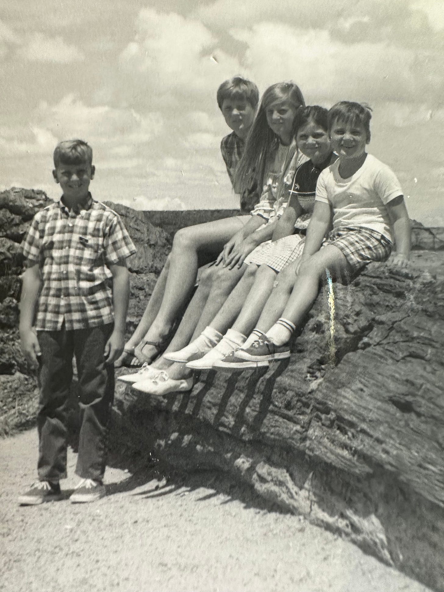 A balcka nd white photo of five siblings at the Petrified Forest in Arizona. One boy standing; three girls and a boy sitting atop a giant, fallen petrified tree trunk.