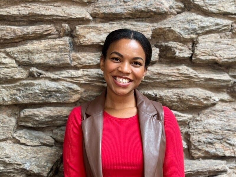 A Black woman who appears to be in her 20s or 30s with a bright smile stands in front of a textured stone wall. She is wearing a red top and a brown leather vest, and her hair is tied back.