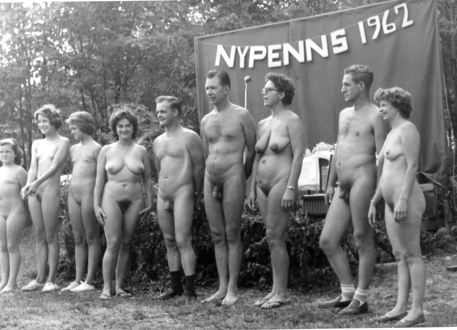 A group of nude adults standing in a row outdoors in front of a banner reading “NYPENNS 1962.”