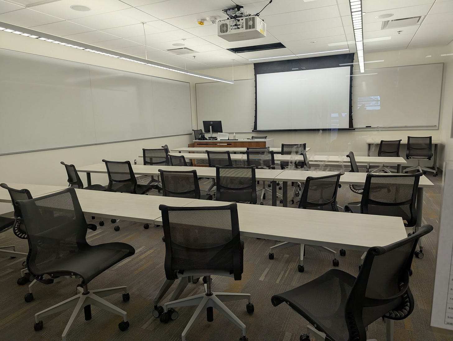 A classroom with long tables, rolling chairs, whiteboards on two walls, and a projector at the front.