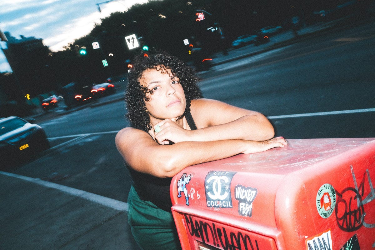 Image of a woman with black curly hair leaning against an orange newspaper dispenser covered in stickers and graffiti on a city sidwalk