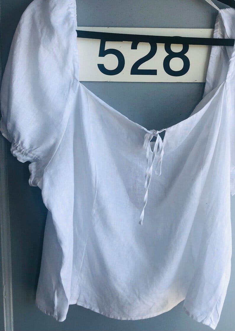 May include: A white square-neck top with short, puffed sleeves and a tie detail at the neckline. The number "528" is visible on a sign in the background. The top is made of a lightweight fabric.