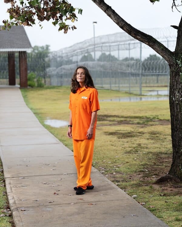 A dark-haired woman wearing an orange shirt and orange pants standing on a sidewalk with fencing behind her. A dark-haired woman wearing an orange shirt and orange pants standing on a sidewalk with fencing behind her.