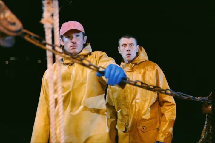 Two men in yellow fisherman's slickers stare at something off camera. A chain pulley system is in the foreground of the shot.