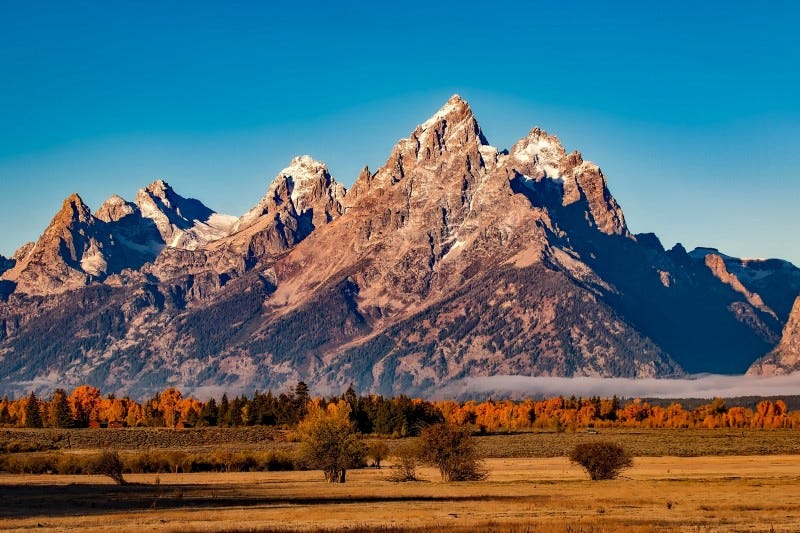 Mountains with snowy peaks. Autumn forest below them, with a thin wisp of fog rolling over the tips. Mountains with snowy peaks. Autumn forest below them, with a thin wisp of fog rolling over the tips.