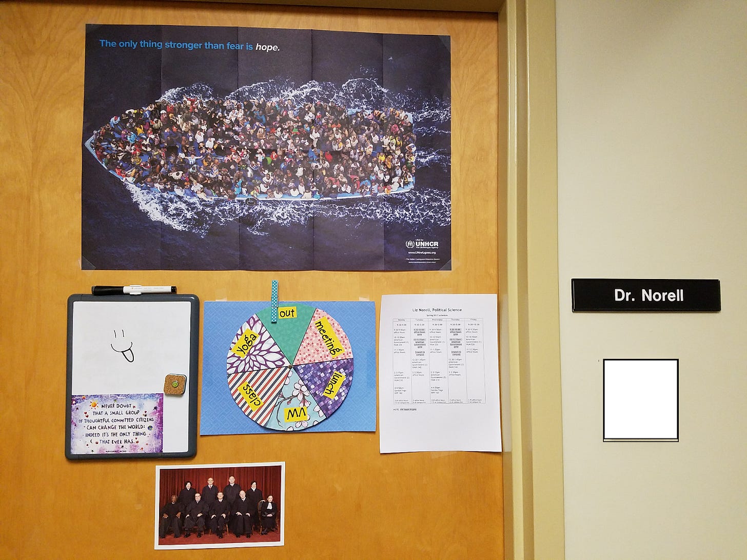 a brown wooden door is decorated with an UNHCR poster, a whiteboard on which someone has drawn a face with the tongue sticking out, a colorful spinning wheel indicating where the office occupant is (currently on out), a door schedule, and a picture of the Supreme Court justices. To the right is a nameplate that says “Dr. Norell.”