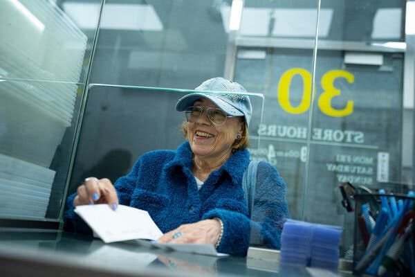 Jennifer Tessler, in a blue cap and blue fleece jacket, receives a check for $7,338 after selling gold baby charms and jewelry at Bullion Exchanges, a gold trading store on 47th Street in the diamond district of Manhattan.