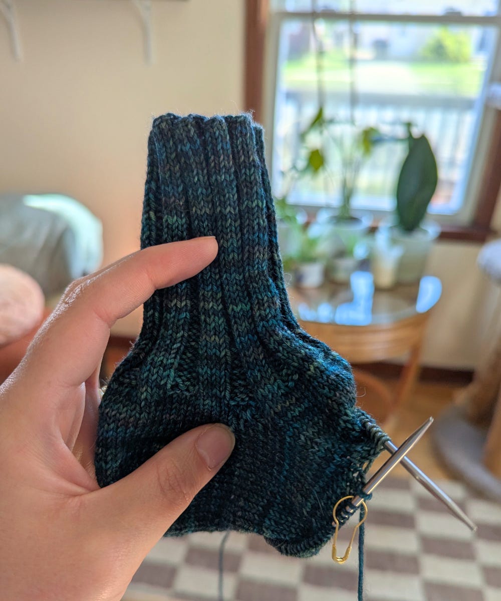 A hand holds a knitted project: a sock in progress. The cuff and heel are complete, two knitting needles stick out of one end.