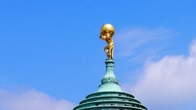 Golden statue of Atlas struggling under weight at the top of a copper building roof. Golden statue of Atlas struggling under weight at the top of a copper building roof.