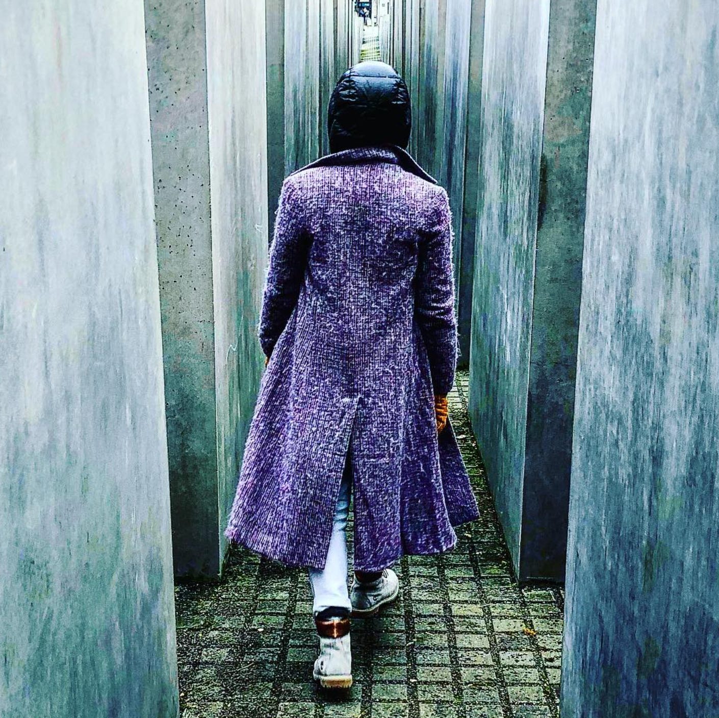 Person in a long purple coat walking through narrow concrete slabs of the Memorial to the Murdered Jews of Europe in Berlin.