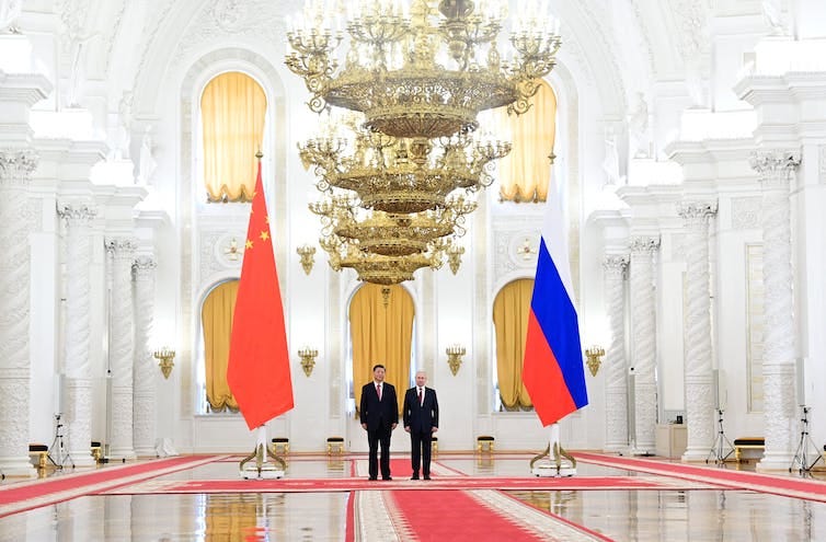 Two men are seen in the background flanked by giant China and Russian flags. Chandeliers hang overhead. Two men are seen in the background flanked by giant China and Russian flags. Chandeliers hang overhead.