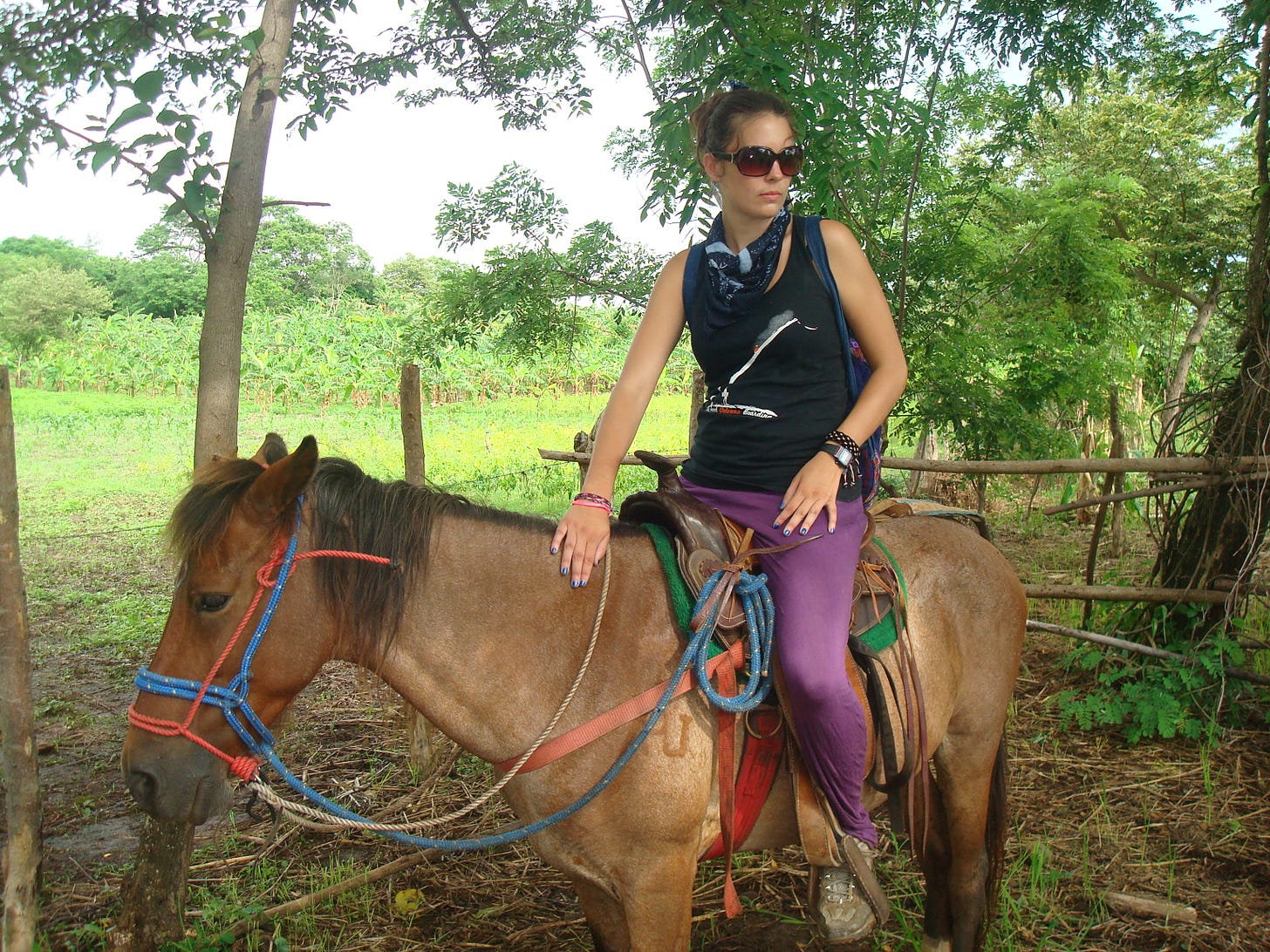 Caitlin, wearing big sunglasses, purple pants and a black tank top sits on a brown horse.