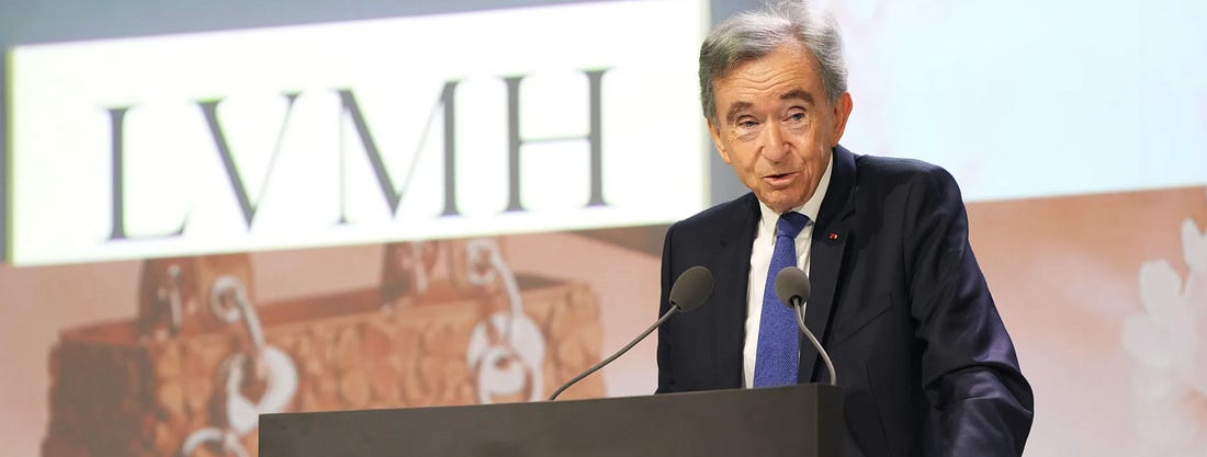 Bernard Arnault, Chairman and CEO of LVMH, speaking at a podium with two microphones in front of a backdrop displaying the LVMH logo and Louis Vuitton monogram trunks during the group's 2026 Annual General Meeting at the Carrousel du Louvre on April 23rd, where he addressed shareholders on a 5% revenue decline to €80.8bn, the Middle East crisis impact on Q1 growth, and succession questions involving his five children.