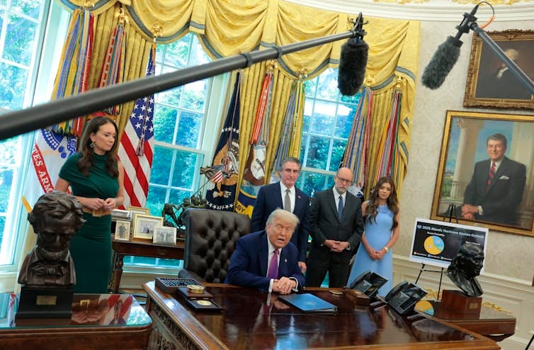 An older white man with a navy-blue suit sits at a large wooden table and is surrounded by four people dressed formally in an ornate room. Large microphones are seen hanging above the desk. An older white man with a navy-blue suit sits at a large wooden table and is surrounded by four people dressed formally in an ornate room. Large microphones are seen hanging above the desk.