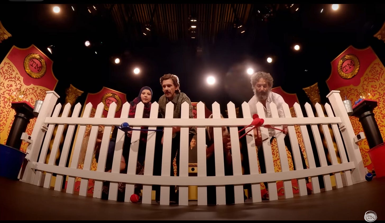 Taskmaster contestants on stage behind a white picket fence, using long-handled wooden spoons to pass tennis balls through the slats in the fence.