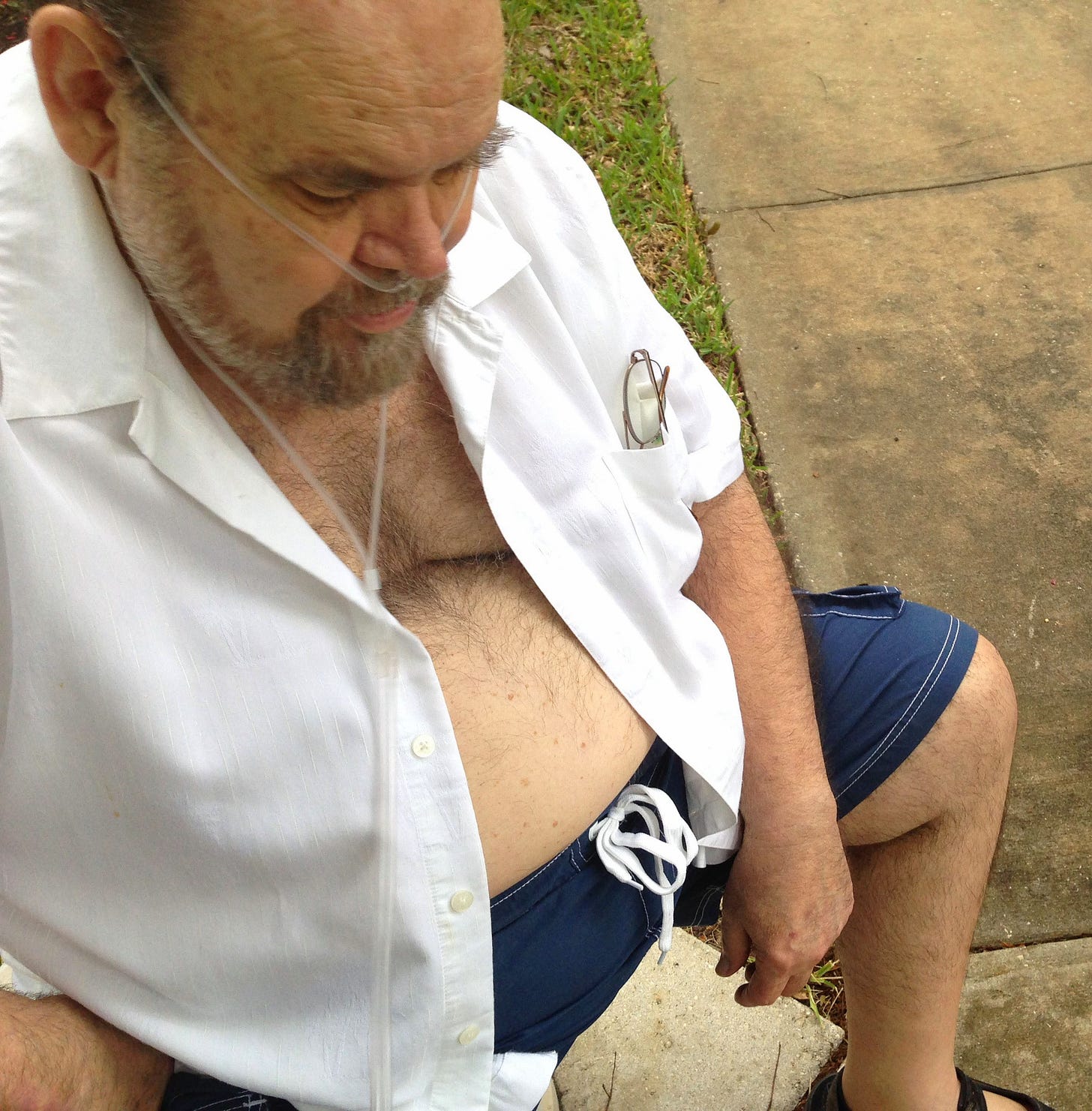 An elderly man sits, looking down, with oxygen tubes in his nose. He is wearing blue swim trunks and an open button front shirt.