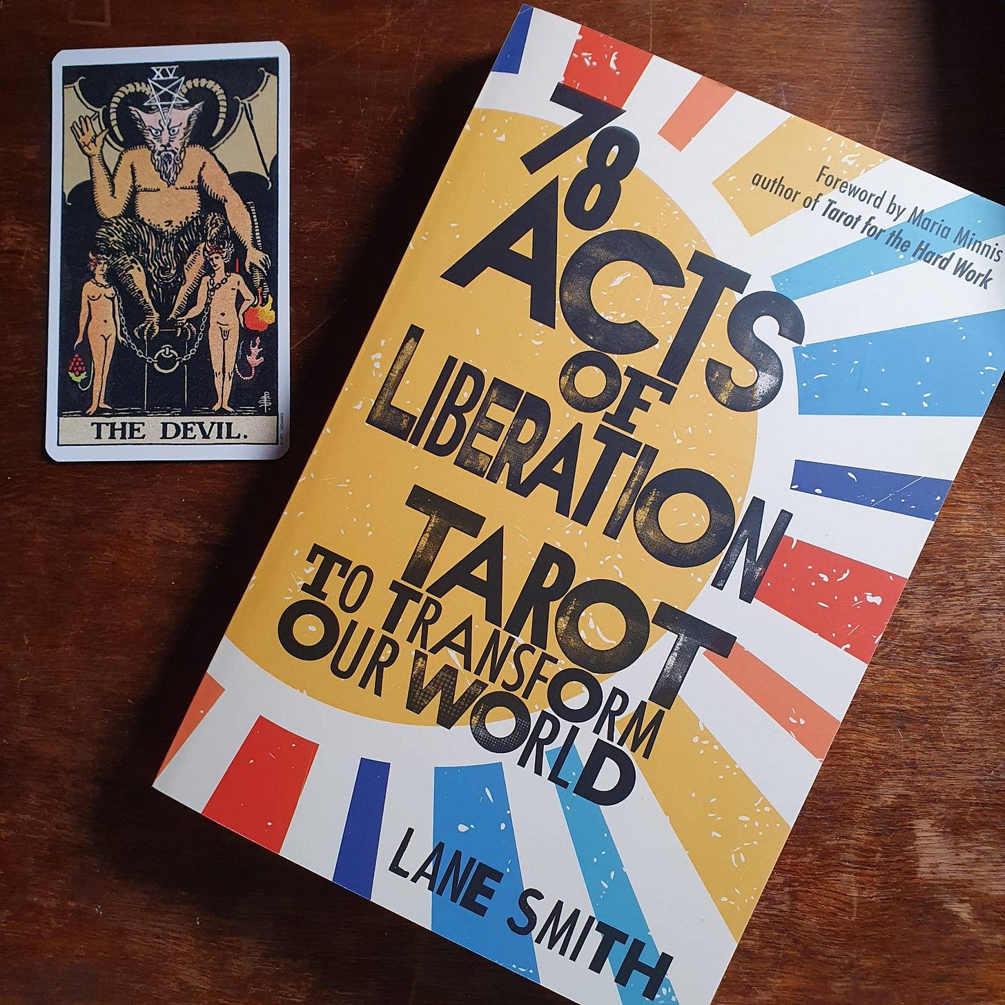 The book "78 Acts of Liberation Tarot to Transform Our World" on a desk beside The Devil tarot card from the Rider Waite Smith deck