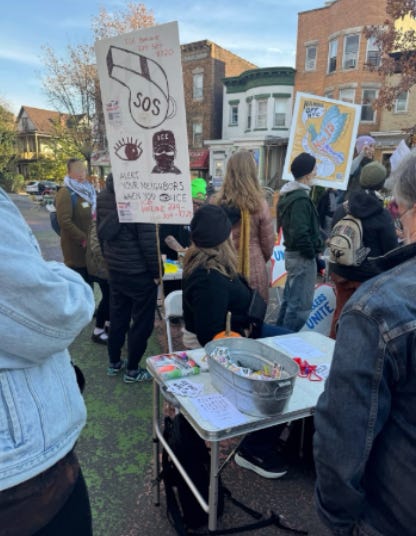photo of people outside in a crowd. There is a sign with an illustration of a whistle and the letters SOS with an eye and an image of a man in an ice hat. One woman is standing in front of a table with whistles and markers