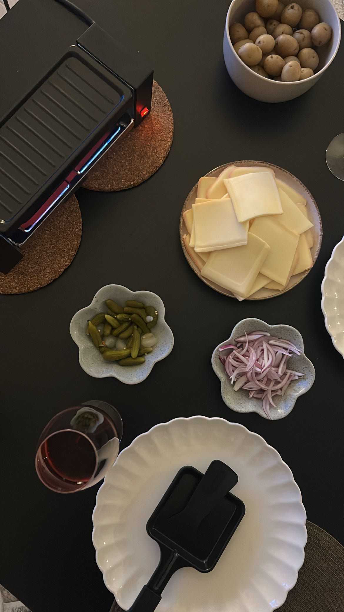 Overhead view of a raclette dinner on a dark table: an electric raclette grill on cork mats, a plate stacked with square slices of raclette cheese (some with brown smoked rinds), small flower-shaped dishes of cornichons, pickled onions and sliced shallots, a glass of red wine, and a black raclette pan resting on a white scalloped plate.