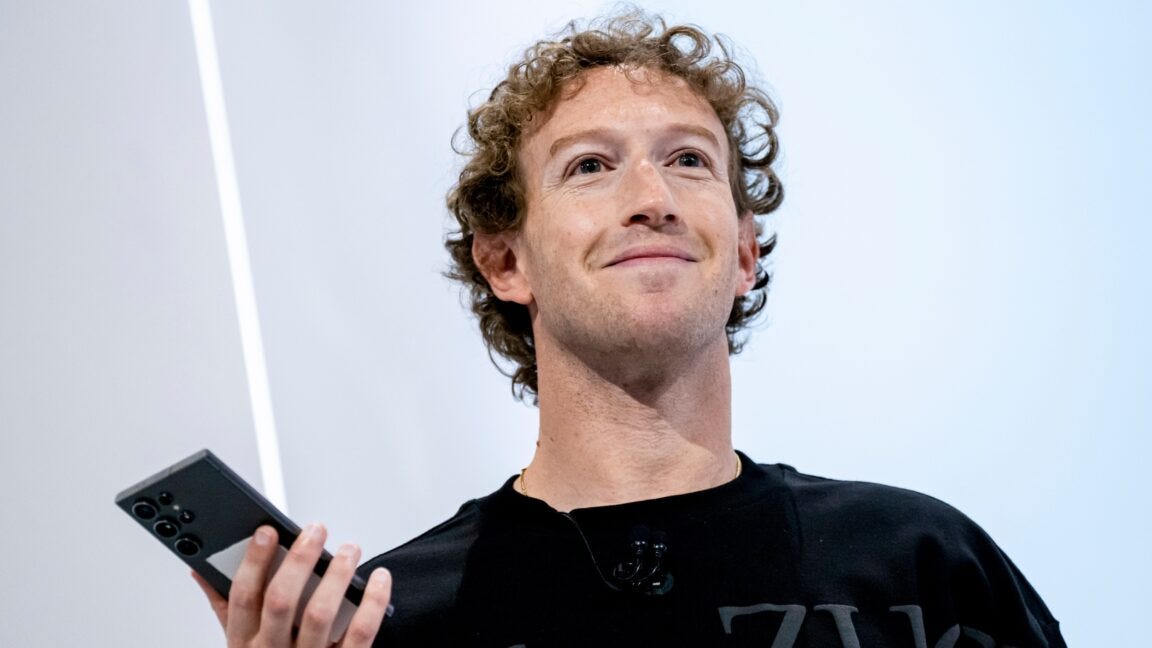 Meta CEO Mark Zuckerberg standing on stage during an event, smiling, and holding a phone in his right hand.