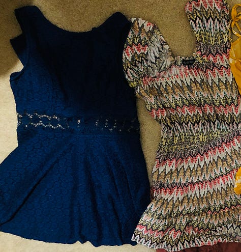 A pile of floral dresses teeters on the thrift store bench. Four are chosen: a goldenrod, a white-with-purple, a midnight blue with a lace cutout at the waistband, and a multi-hued jaggedy pattern with puffy cap sleeves.
