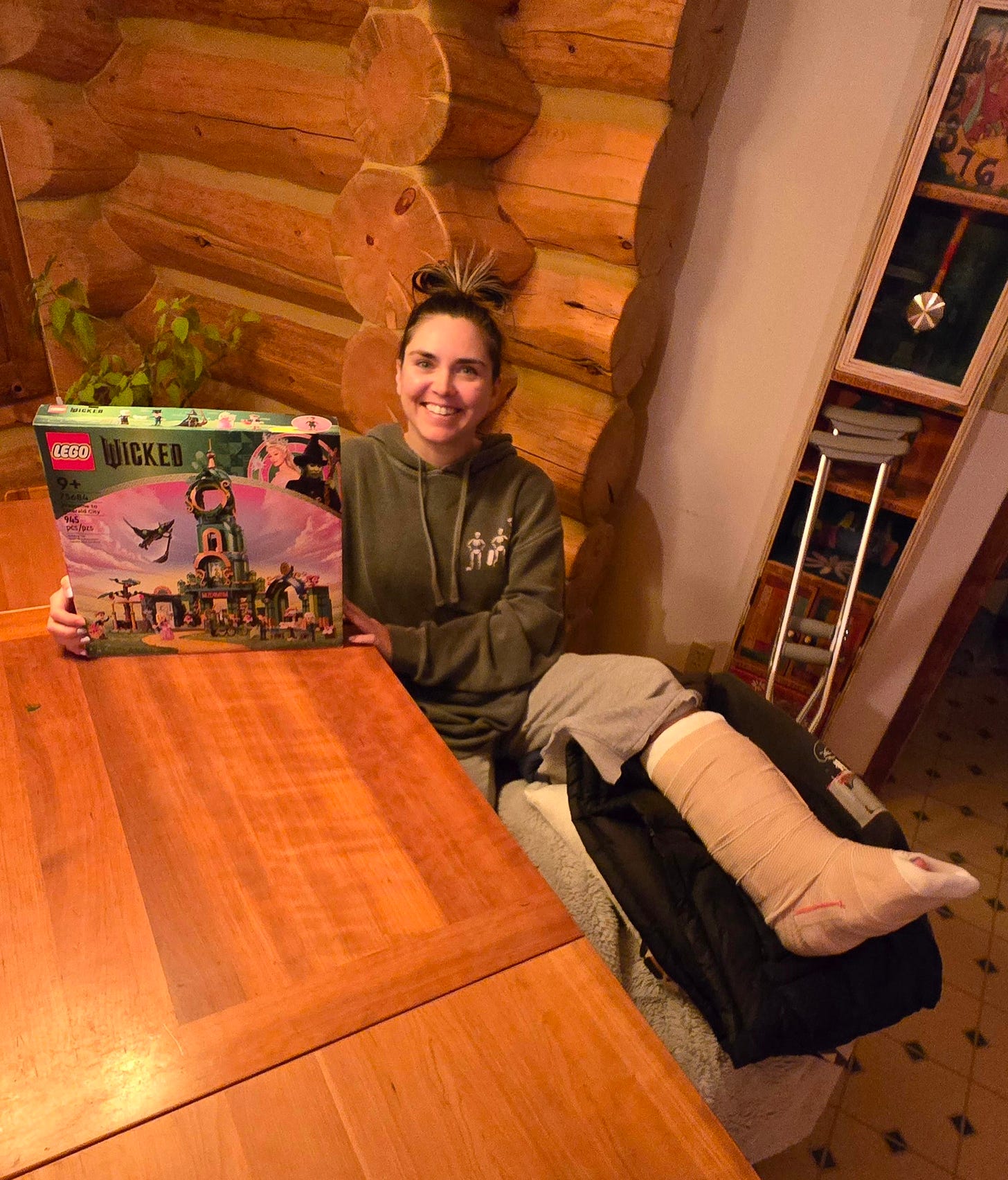 Cassidy sitting at a table with her broken leg propped up on a chair, holding a brand new Wicked Lego set in the box