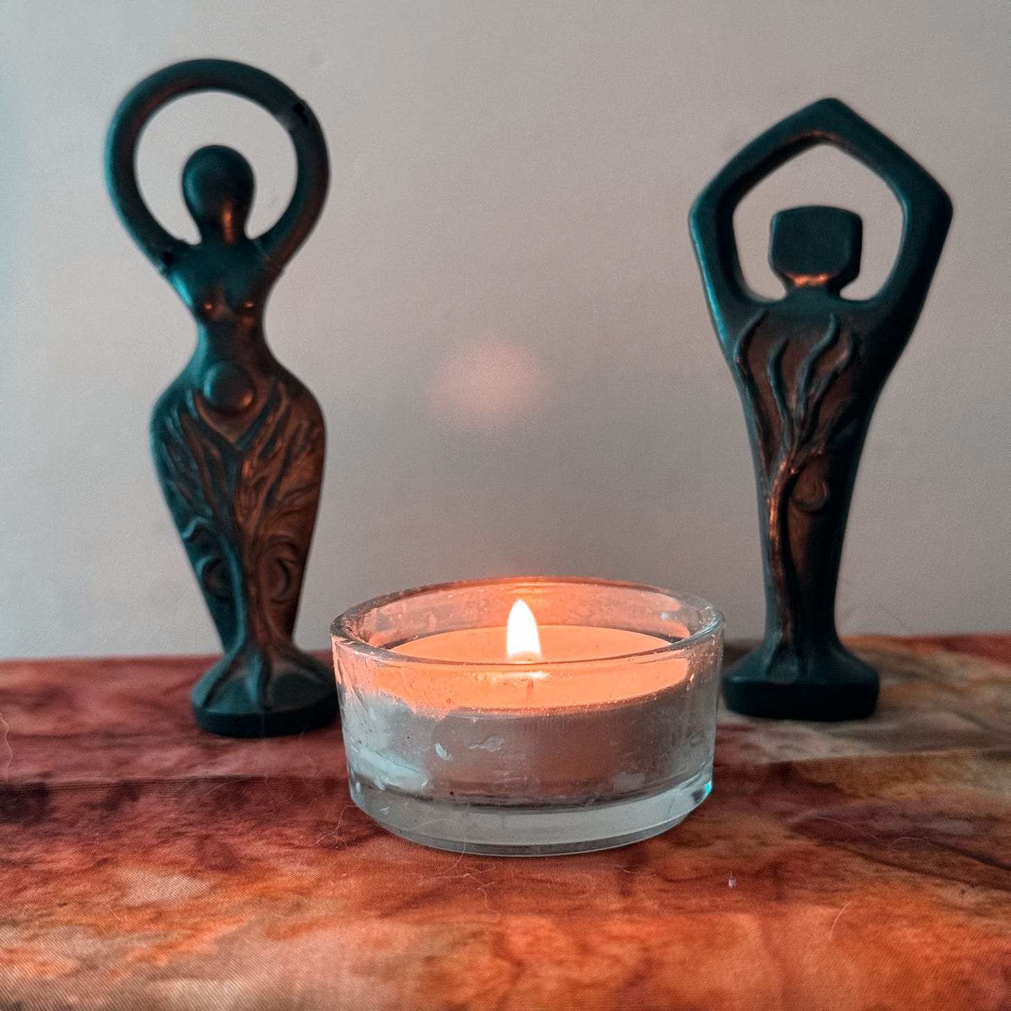 A pair of black statuettes stand on an autumnal-coloured cloth with a lit tealight candle between them and slightly to the front. The statuette on the left shows a curving female silhouette with her arms linked in a circle above her head. The statuette on the right shows a male silhouette in the same pose.