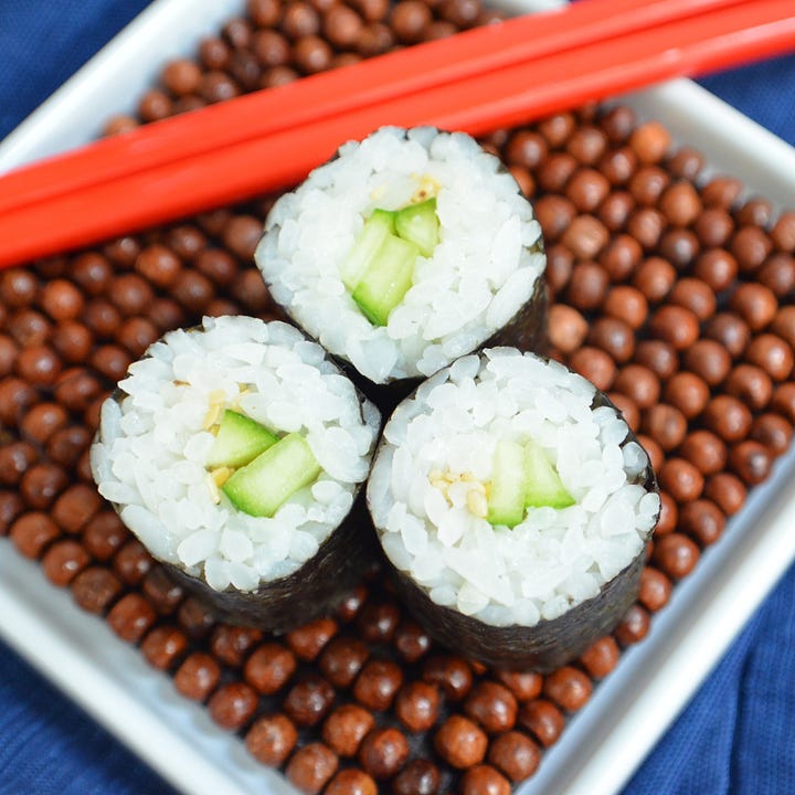 Two types of hosomaki sushi rolls. One is filled with roasted butternut squash and candied ginger; the other is a classic cucumber roll topped with sesame seeds. Both are sliced into bite-sized pieces and arranged on a wood plate.