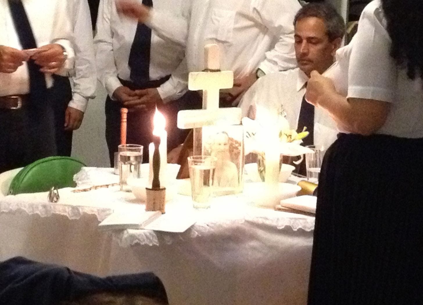 Candlelit table with a white cloth, water glasses, a two-bar wooden cross and a framed photo of a bearded man, men in white shirts and navy ties standing behind, faces out of frame. Candlelit table with a white cloth, water glasses, a two-bar wooden cross and a framed photo of a bearded man, men in white shirts and navy ties standing behind, faces out of frame.