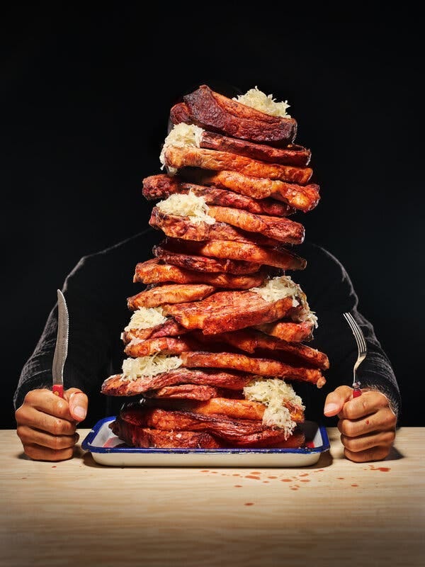 A man sits behind a plate of food, a fork and knife clenched in his fists. On the plate is a stack of grilled meats and sauerkraut so tall it obscures his face. Meat juice is splattered on the table.